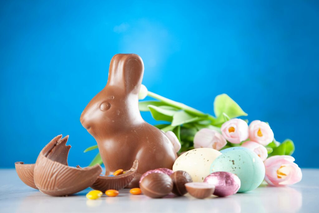A delightful Easter scene with a chocolate bunny, colorful eggs, and pink tulips on a blue background.